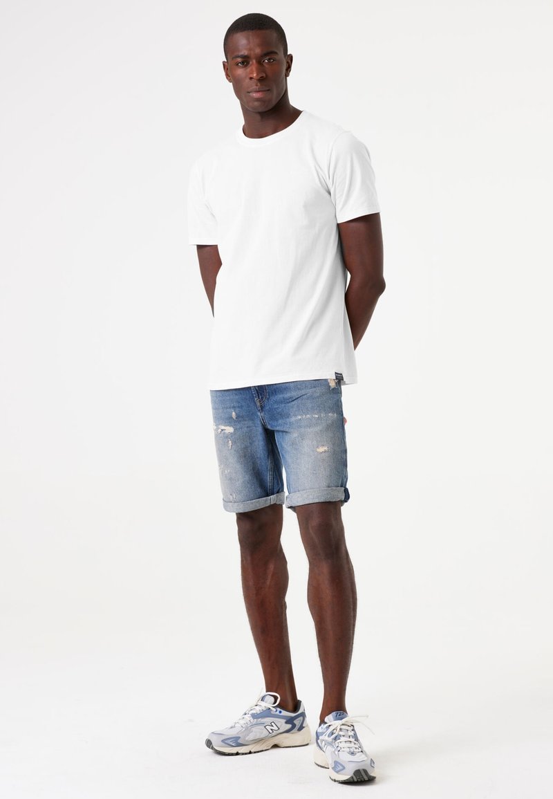 Camiseta blanca de manga corta combinada con shorts de denim azul desgastados, con dobladillos enrollados y zapatillas grises con acentos blancos.