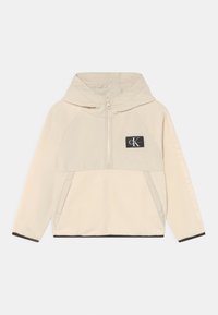 Pullover beige con cappuccio realizzato in morbido pile, presenta una zip frontale, due tasche laterali e un patch con logo. Rifinito in nero all'orlo.