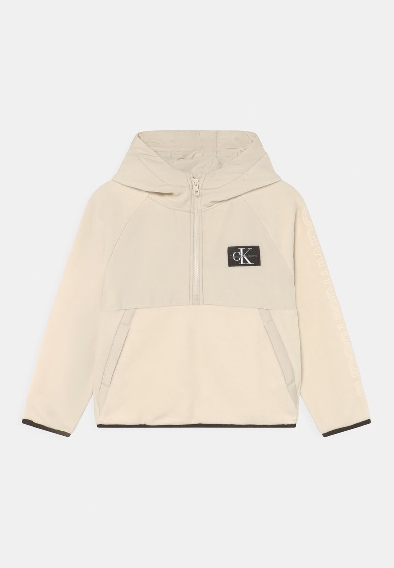 Pullover beige con cappuccio realizzato in morbido pile, presenta una zip frontale, due tasche laterali e un patch con logo. Rifinito in nero all'orlo.