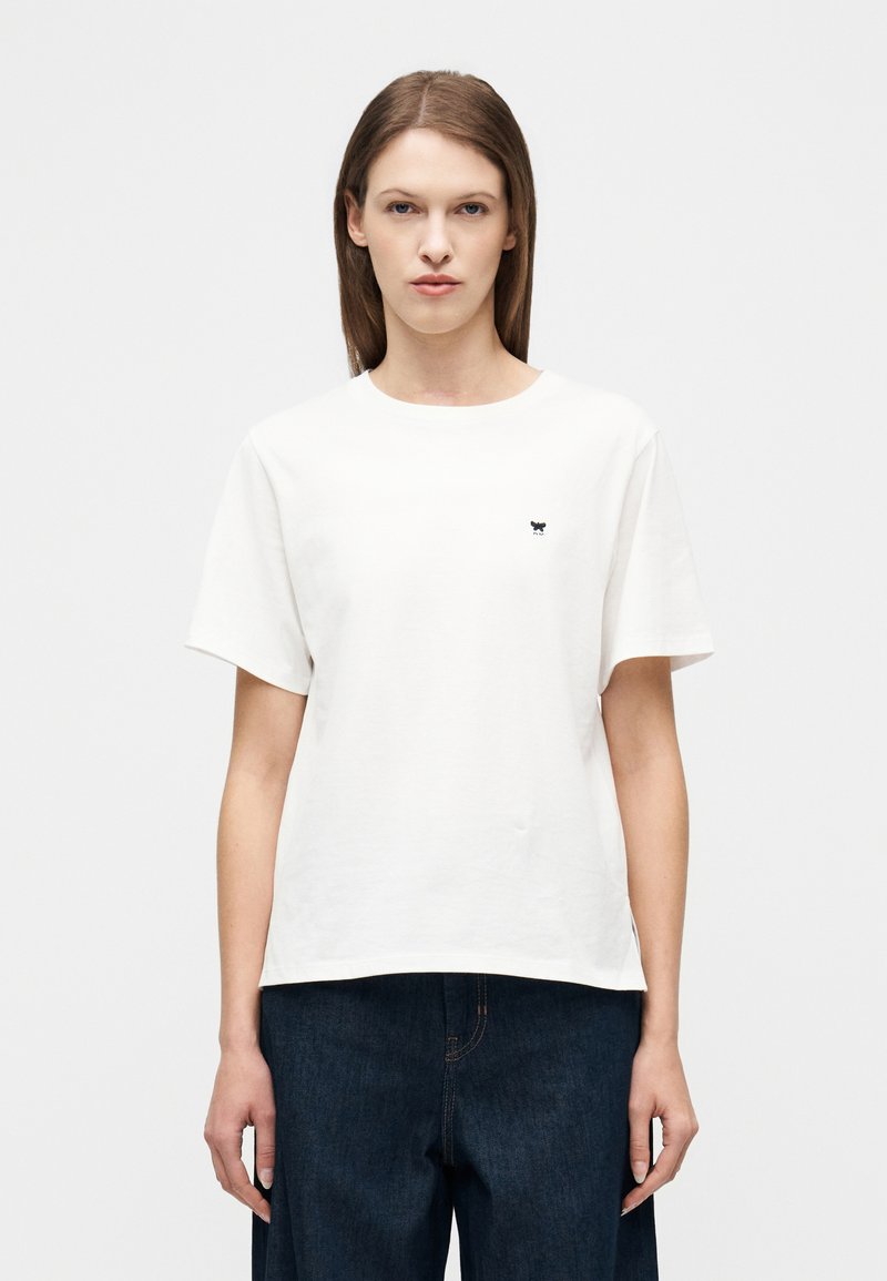 Γυναίκα με ίσια καστανά μαλλιά φορ άει λευκό κοντομάνικο μπλουζάκι και σκούρο μπλε παντελόνι, στέκεται μπροστά σε ένα απλό φωτεινό φόντο.