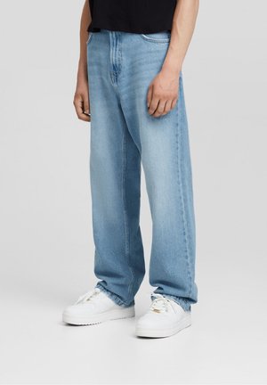 Person iført lyseblå vidde jeans, hvide sneakers og en sort skjorte, stående mod en ensartet lys baggrund.