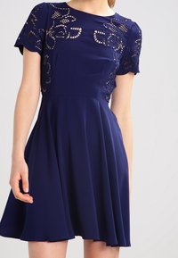 Navy blue dress with short sleeves, featuring intricate lace cutouts and a fitted bodice that flares into a flowing skirt.