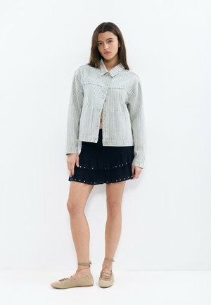 Mujer joven con chaqueta clara de rayas y botones, mini falda negra de volantes con tachuelas y zapatos planos beige con cordones, sobre un fondo blanco.