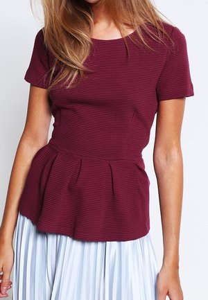 Nainen, joka pukeutunut istuvaan, lyhythihaiseen viininpunaiseen toppiin, jossa on tekstuurilliset vaakasuorat raidat ja laskostettu peplum-vyötärö, yhdistettynä vaaleaan laskostettuun hameeseen.