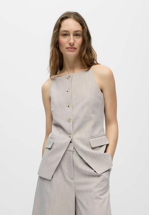 Femme aux longs cheveux portant un gilet beige sans manches boutonné et un pantalon large assorti, debout les mains dans les poches.