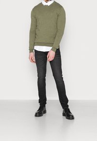 Maglione verde oliva con collo rotondo, abbinato a una camicia bianca con colletto, jeans neri slim fit e stivali neri stringati. Tessuto morbido.