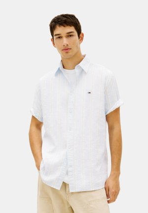 Ung mand iført en hvid kortærmet skjorte over en hvid t-shirt og beige bukser, stående med den ene hånd i lommen.