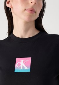 Black cotton t-shirt featuring a square logo in pink and teal with "cK" in white text. Round neckline and short sleeves.