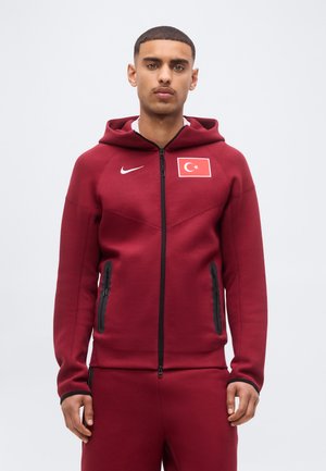 Młody mężczyzna w bordowej bluzie Nike na zamek i dopasowanych spodniach z naszywką tureckiej flagi na lewej piersi.