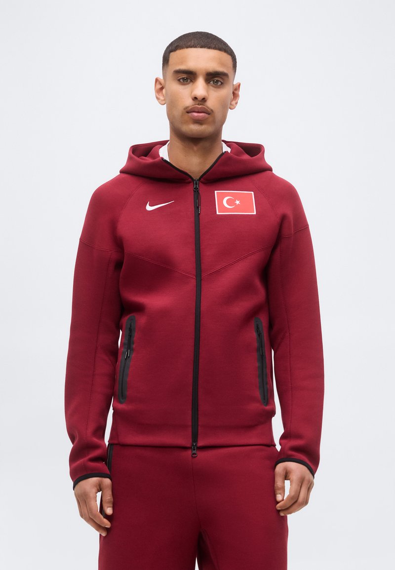 Νέος άνδρας φοράει μπορντό φούτερ Nike με φερμουάρ και ταιριαστό παντελόνι με σήμα της τουρκικής σημαίας στο αριστερό στήθος.