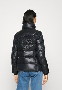 Kvinna som bär en svart, glänsande puffjacka och blå jeans, vänd bort från kameran mot en ljus vit bakgrund.
