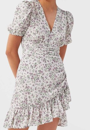 Witte bloemen wikkeljurk met pofmouwen, gerimpelde taille en geruite zoom gedragen door een persoon tegen een effen achtergrond.