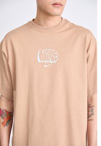 Bež oversized majica od pamuka s svijetloplavim grafičkim logotipom na prsima; kratki rukavi; glatka tekstura; minimalistički dizajn.