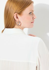 Camicia bianca con schiena plissettata, dotata di colletto classico. Il modello indossa un orecchino ad anello in argento lucido. Tessuto a trama liscia.