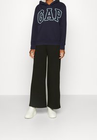 Sweat à capuche Navy GAP avec de grandes lettres bleu clair, associé à un pantalon large noir et des baskets blanches. Tenue décontractée et confortable.