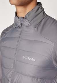 Pilkas izoliacinis švarkas su siuvinėtu dizainu, aukštu krageliu ir užtrauktuku. Ant krūtinės yra sidabrinis „Columbia“ logotipas. Minkšta, lengva medžiaga.
