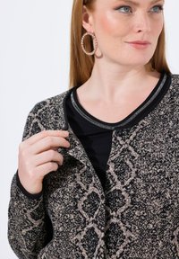 Sweter kardigan w czerni i beżu z teksturowanym, geometrycznym wzorem, z dekoltem w kształcie litery V i zapięciem na guziki. Nosi się go na czarnej bluzce.