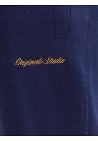 Teddy modra bombažna tkanina z gladko teksturo; vsebuje zlato vezenje z napisom "Originals Studio" na levi strani blizu žepa.