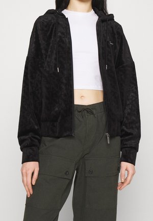 Black zip-up velour hoodie with geometric pattern, drawstring hood, and silver hardware accents, paired with olive green cargo pants.