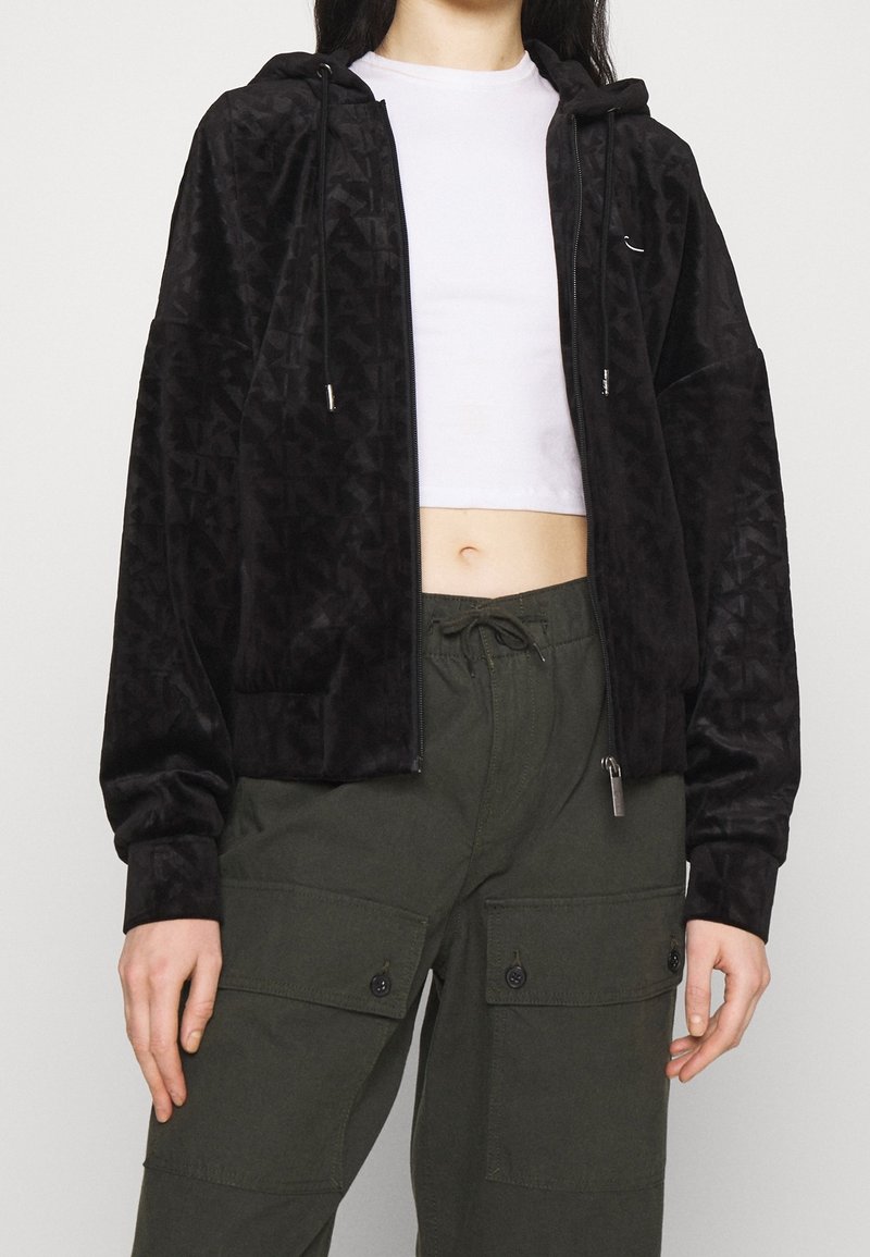 Felpa nera in velluto con zip, motivo geometrico, cappuccio con cordino e rifiniture in metallo argentato, abbinata a pantaloni cargo verde oliva.