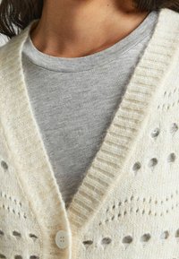 Krämbelagd stickad cardigan med djup V-ringning, featuring knapplås och öppet arbete detaljer. Grå crewneck-tröja synlig under.