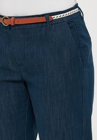 Un pantalon en denim bleu foncé présente des poches avant, une ceinture en cuir marron avec une boucle en or et un accent tressé blanc et marron.