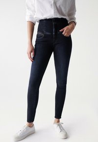 Žena v tmavých skinny džínách s vysokým pasem, bílé košili s dlouhým rukávem a ohrnutými rukávy, a bílých teniskách, stojící s jednou rukou v kapse.