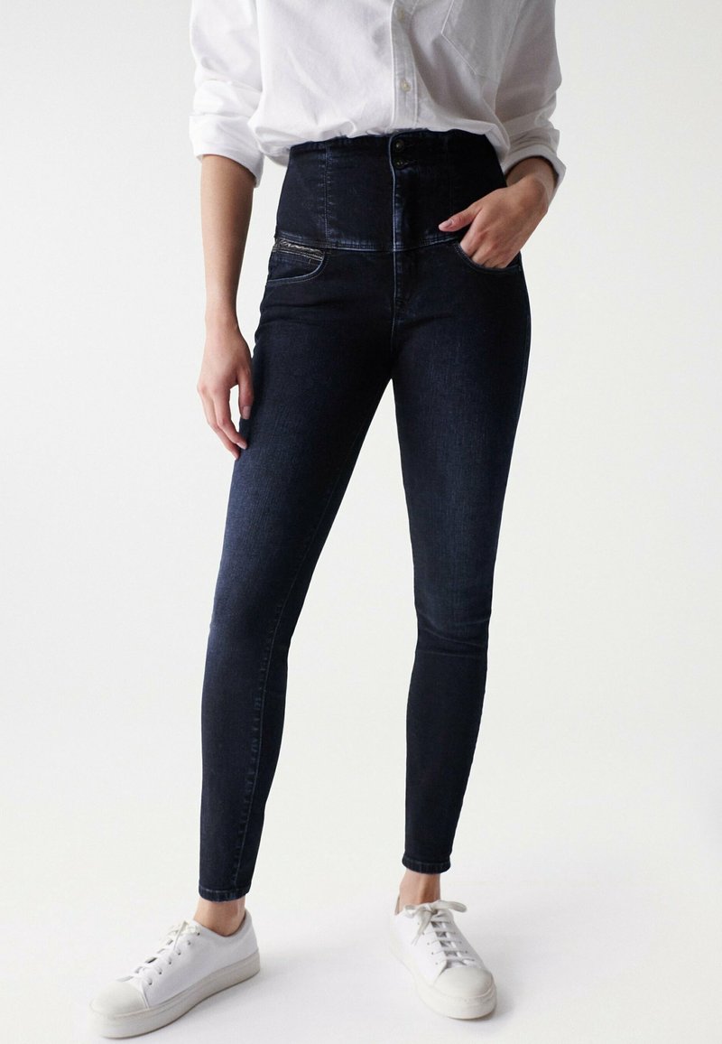 Žena v tmavých skinny džínách s vysokým pasem, bílé košili s dlouhým rukávem a ohrnutými rukávy, a bílých teniskách, stojící s jednou rukou v kapse.