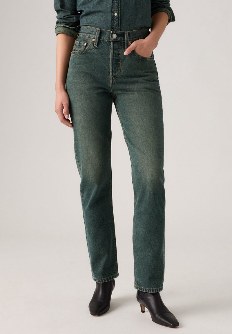 Personne portant un jean taille haute vert foncé avec une chemise en denim rentrée, une main dans la poche et des bottines à talons noires.
