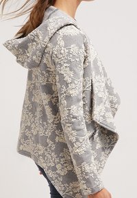 Giacca con cappuccio grigia a motivo floreale con finitura texturizzata. Presenta un frontale drappeggiato e un retro più lungo, accentuato da ricami intricati.
