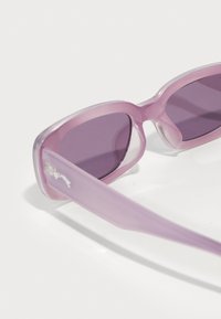 Lunettes de soleil transparentes violettes avec des verres teintés, un design épuré et anguleux, et une finition lisse. Les branches sont légèrement translucides avec un détail de logo.