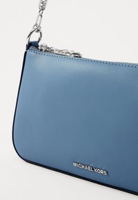 Svijetloplava kožna torba preko tijela sa srebrnim detaljima, glatke teksture, zatvaranje na patentni zatvarač i urezanim logotipom "MICHAEL KORS" sprijeda.