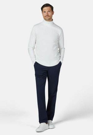 Man staat met handen in de zakken, draagt een witte coltrui, marineblauwe broek en witte sneakers op een effen lichte achtergrond.