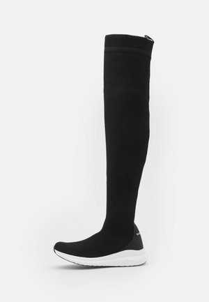 Bottes montantes noires en maille stretch avec un design élégant, dotées d'une semelle en caoutchouc blanche et d'une tige texturée. Esthétique minimaliste et moderne.