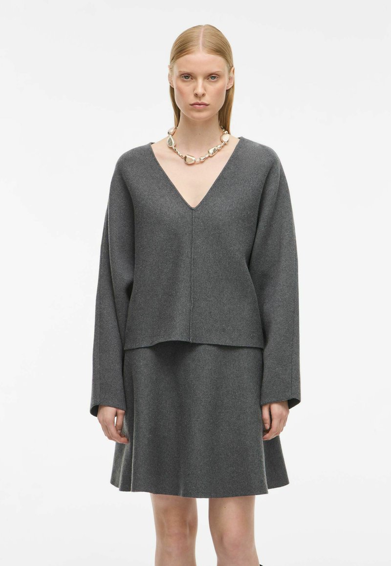 Siv pulover iz volne z V-izrezom in dolgimi rokavi, v kombinaciji s krilom v enaki sivi barvi s širokim krojem. Gladka tekstura, minimalistična zasnova, brez vzorcev.