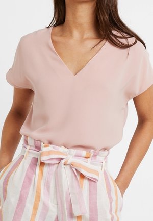 Vrouw die een lichtroze V-hals short-sleeve top draagt, ingestopt in een gestreepte broek met hoge taille en een geknoopte stoffen riem.
