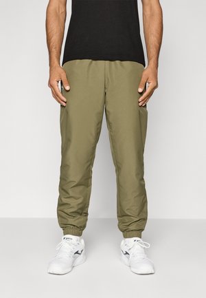 Homme portant un pantalon de jogging vert olive, des baskets blanches et une chemise noire, debout devant un fond clair uni.