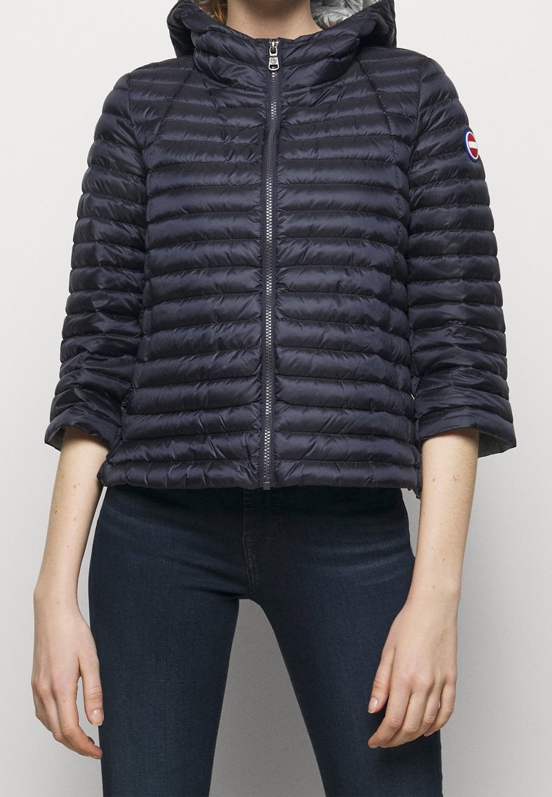 Donna che indossa una giacca imbottita trapuntata blu navy con cerniera frontale e cappuccio, abbinata a jeans aderenti blu scuro.