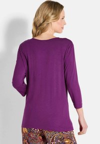 Lila Langarmshirt mit lockerer Passform. Weicher Stoff, runder Ausschnitt und leicht fallende Schultern. Kombiniert mit gemusterten Hosen.