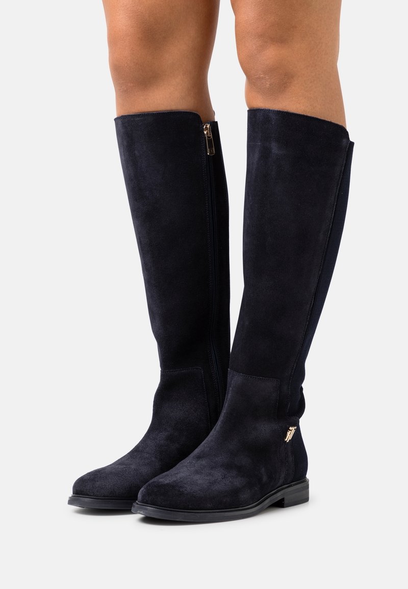 Personne portant des bottes montantes en daim noir avec une fermeture éclair latérale et des talons plats bas, debout sur un fond clair uni.