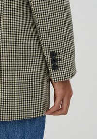 Blazer à motif pied-de-poule avec un schéma de couleurs beige et noir. Présente un revers cranté et trois boutons noirs sur le revers de la manche.