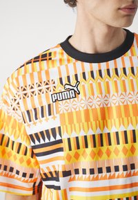 Lyhythihainen, pyöreäkauluksellinen paita, jossa on oranssi, musta ja keltainen geometrinen kuvio, ja PUMA-logo brodeerattuna vasemmassa rinnassa.