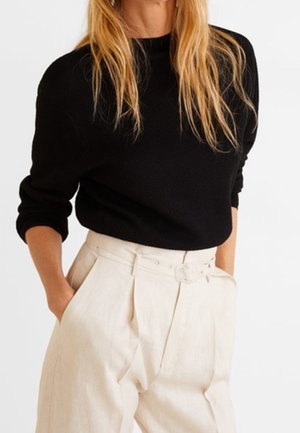 Frau trägt ein schwarzes langärmeliges Oberteil, das in hochtaillierte, cremefarbene Hosen mit Gürtel gesteckt ist, die Hände in den Taschen.