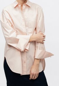 Personne portant une chemise ample rose clair boutonnée, avec des poignets larges non attachés, tenant un bras croisé sur le corps, associée à une jupe foncée.