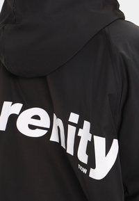 Chaqueta con capucha negra hecha de tejido suave; presenta un gran texto blanco "serenity now" en la parte posterior, enfatizando un diseño moderno y minimalista.