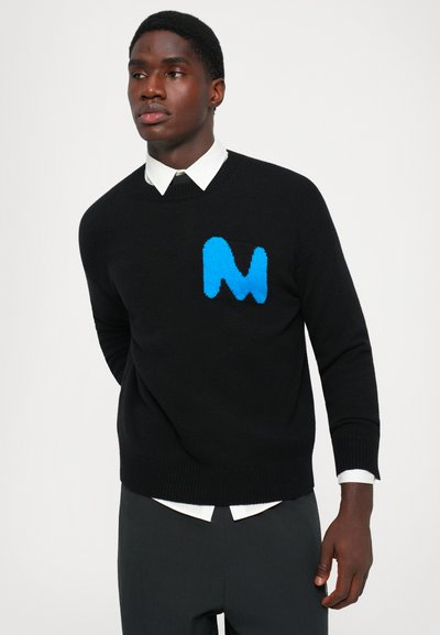 T�ânăr îmbrăcat într-un pulover negru cu un logo albastru "M", cămașă albă cu guler dedesubt și pantaloni închiși, stând în fața unui fundal alb simplu.