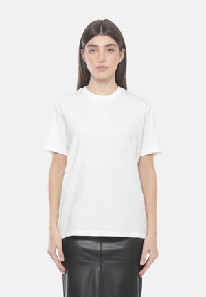 Donna con capelli castani lisci che indossa una t-shirt bianca a maniche corte semplice e una gonna di pelle nera, in piedi davanti a uno sfondo bianco.