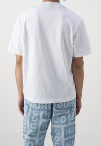 T-shirt bianco in cotone a maniche corte, con un design semplice. Indossato con jeans blu chiaro che mostrano un motivo a logo ripetuto.