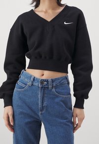 Svart kort sweatshirt med V-hals och ribbad fåll, med en vit Nike swoosh-logotyp. Bärs tillsammans med högmidjade blå jeans.