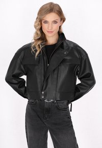 Zwarte leren kortjack met een rits aan de voorkant, een drukknoopsluiting en twee voorzakken. G gedragen met donkergrijze jeans. Gladde textuur.